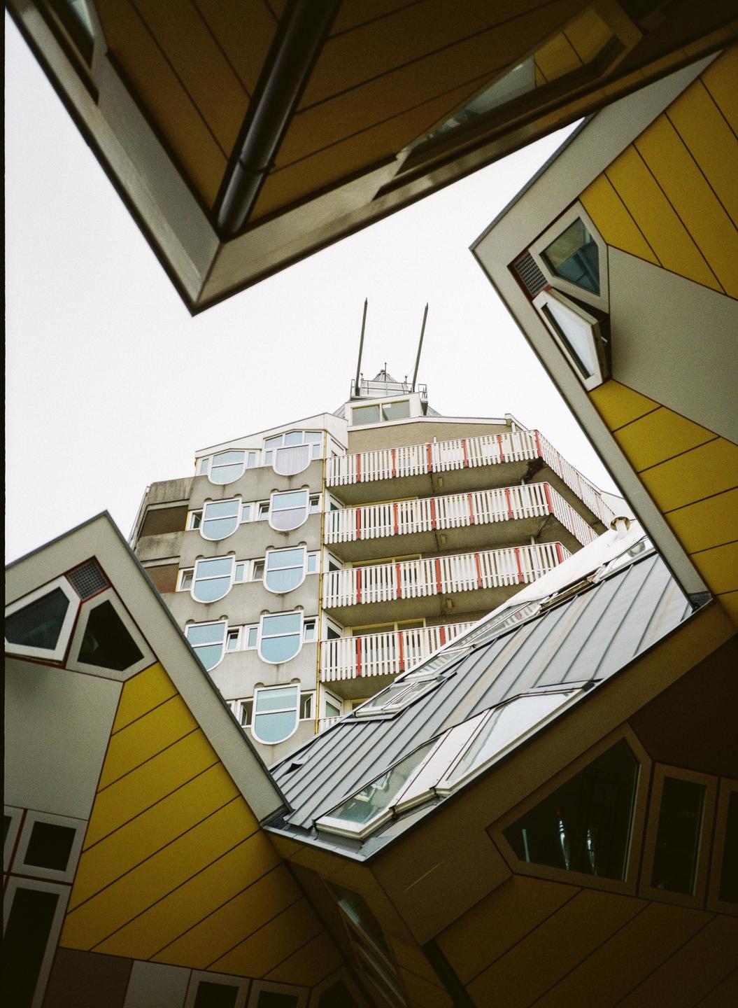 A low-angle view shows the geometric architecture of yellow cube houses framing a residential building with balconies and rounded windows.