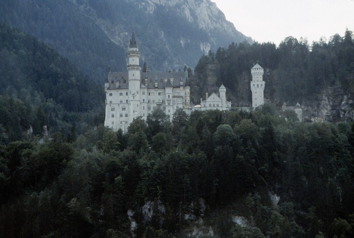 A white castle with multiple towers is nestled among dark green trees on a mountainside.