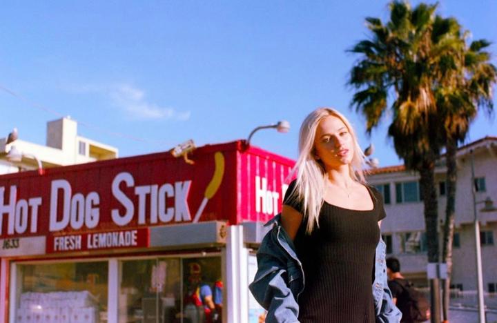 A woman stands in front of a Hot Dog on a Stick stand with palm trees and buildings in the background.