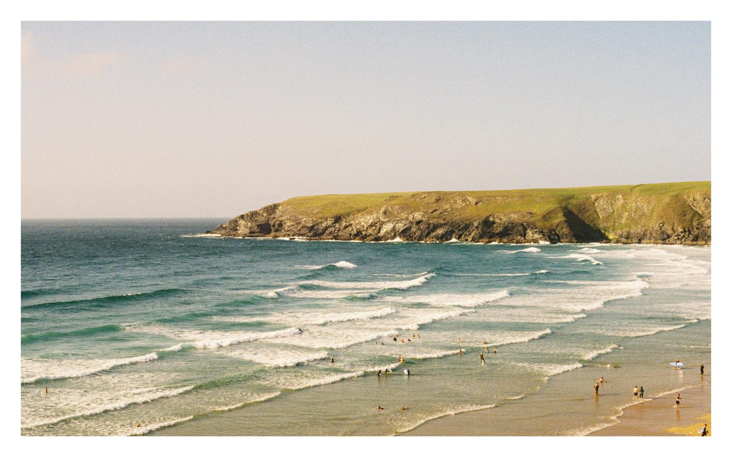 A wide shot shows a beach with many people in the water and on the sand, with a large cliff covered in green grass in the background.