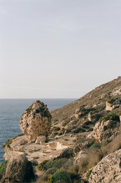 A large rock formation covered in green foliage stands on a cliff overlooking the ocean, with a rocky, vegetated hillside rising to the right.