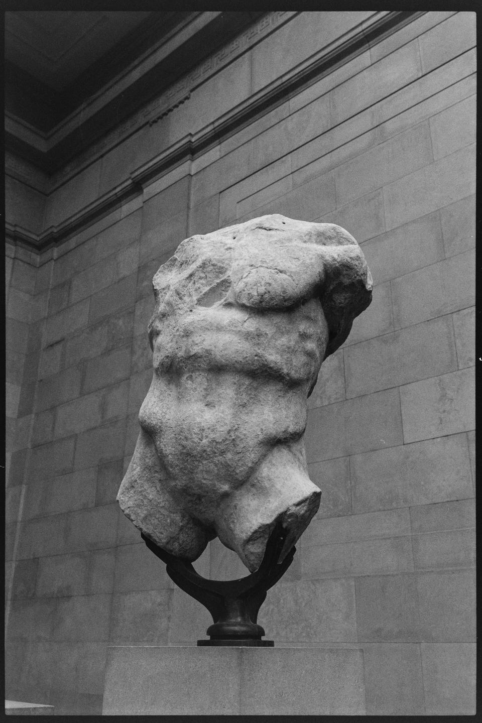 A stone torso sculpture is displayed on a pedestal against a stone wall.