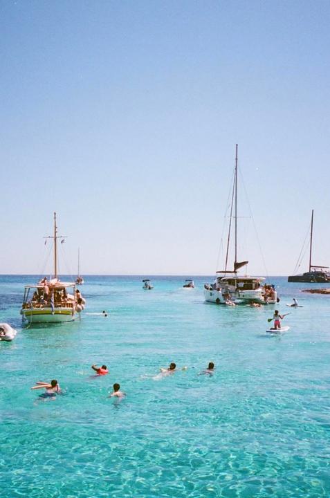 People swim and paddleboard in clear blue water near several boats under a bright sky.