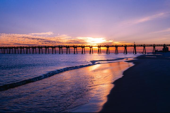A long pier stretches across the water under a colorful sunset sky with waves breaking on the beach.