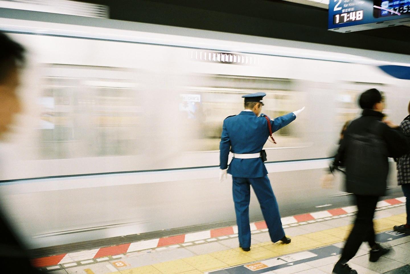 A man in a blue uniform stands on a train platform with a moving train blurring past.
