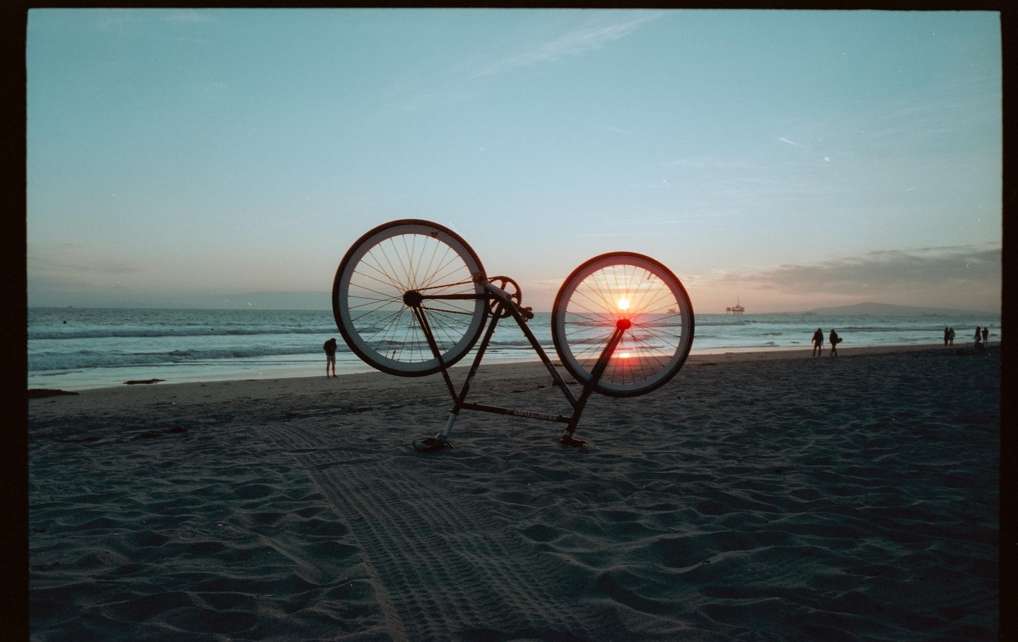A bicycle frame with no tires stands on a sandy beach with the sunset visible through one of its wheels.