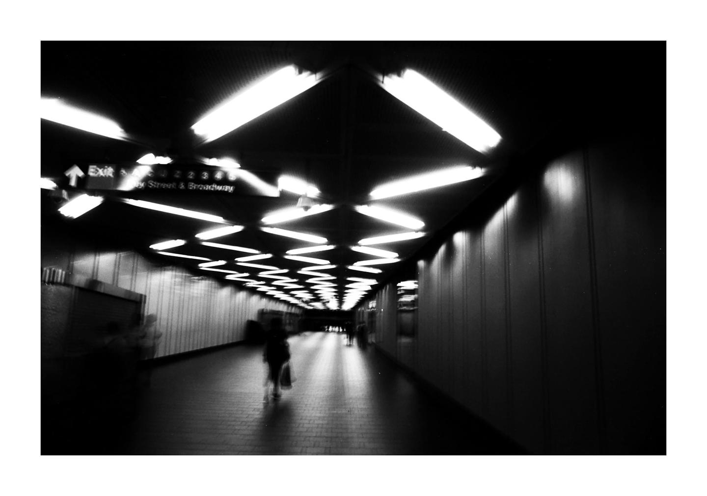 A person walks down a long, brightly lit subway tunnel with an exit sign visible.
