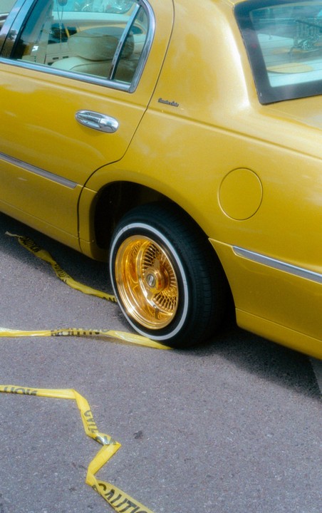 A yellow car with gold rims is parked on asphalt with yellow caution tape on the ground.