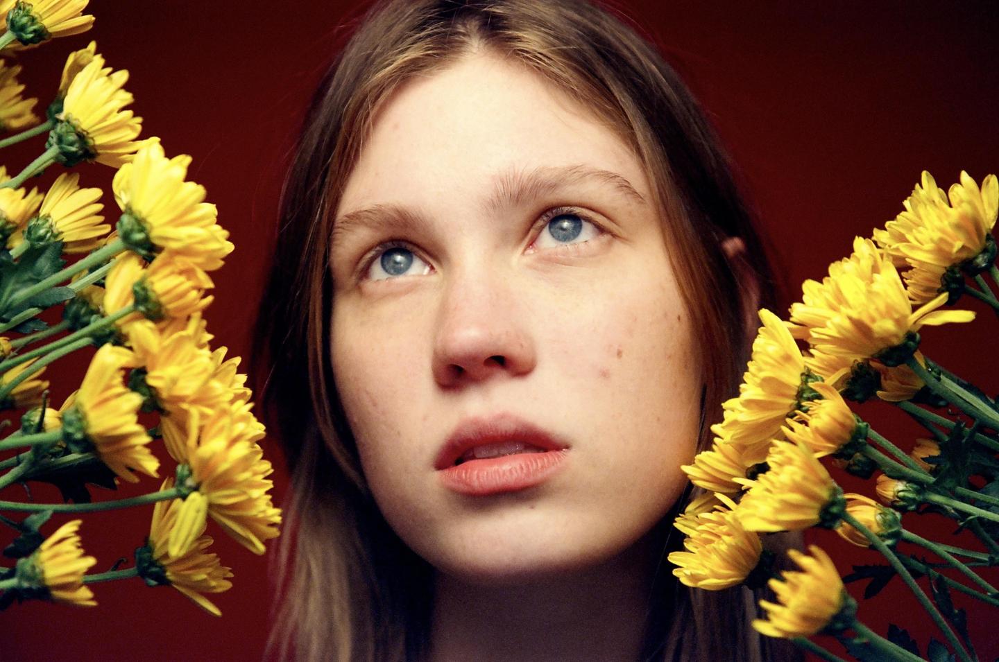A woman with blue eyes looks up, framed by yellow flowers on a red background.