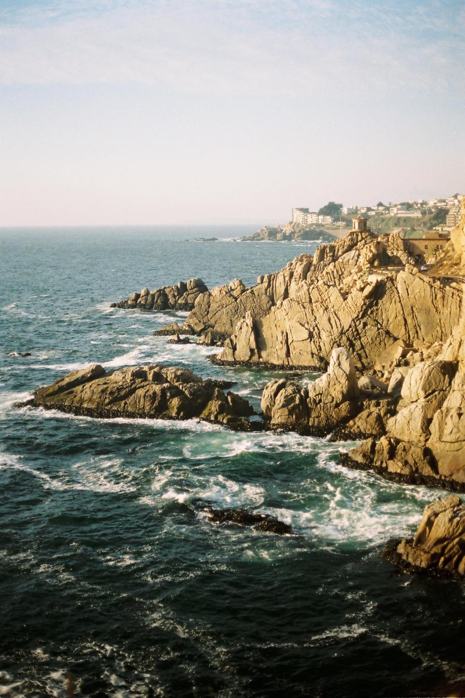 The ocean crashes against a rocky coastline with buildings visible in the distance under a clear sky.