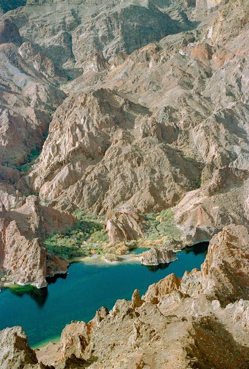 A deep blue lake is nestled among rocky desert mountains with green foliage along its banks.