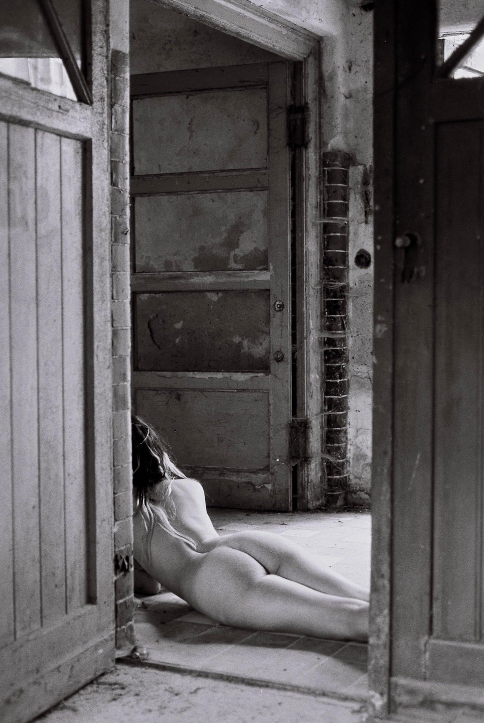 A nude woman lies on the floor of a derelict room, seen from behind.