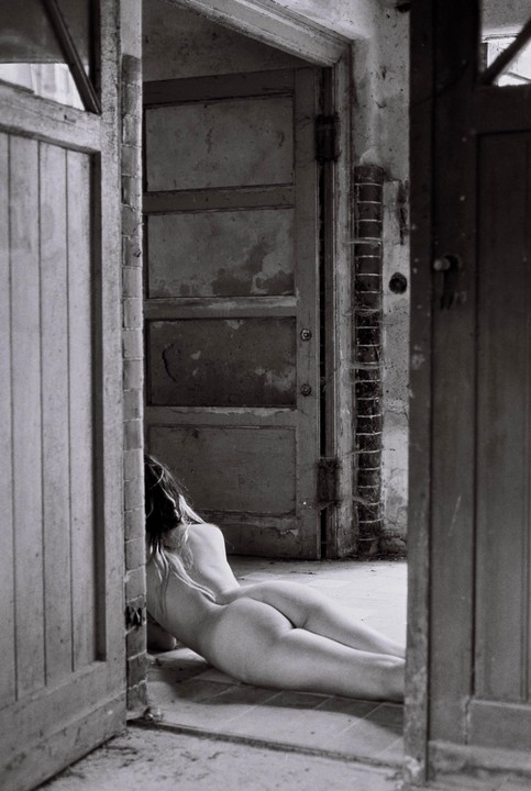 A nude woman lies on the floor of a derelict room, seen from behind.