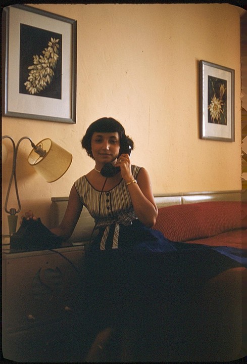 A woman sits on a bed and talks on a telephone next to a lamp and two framed pictures.