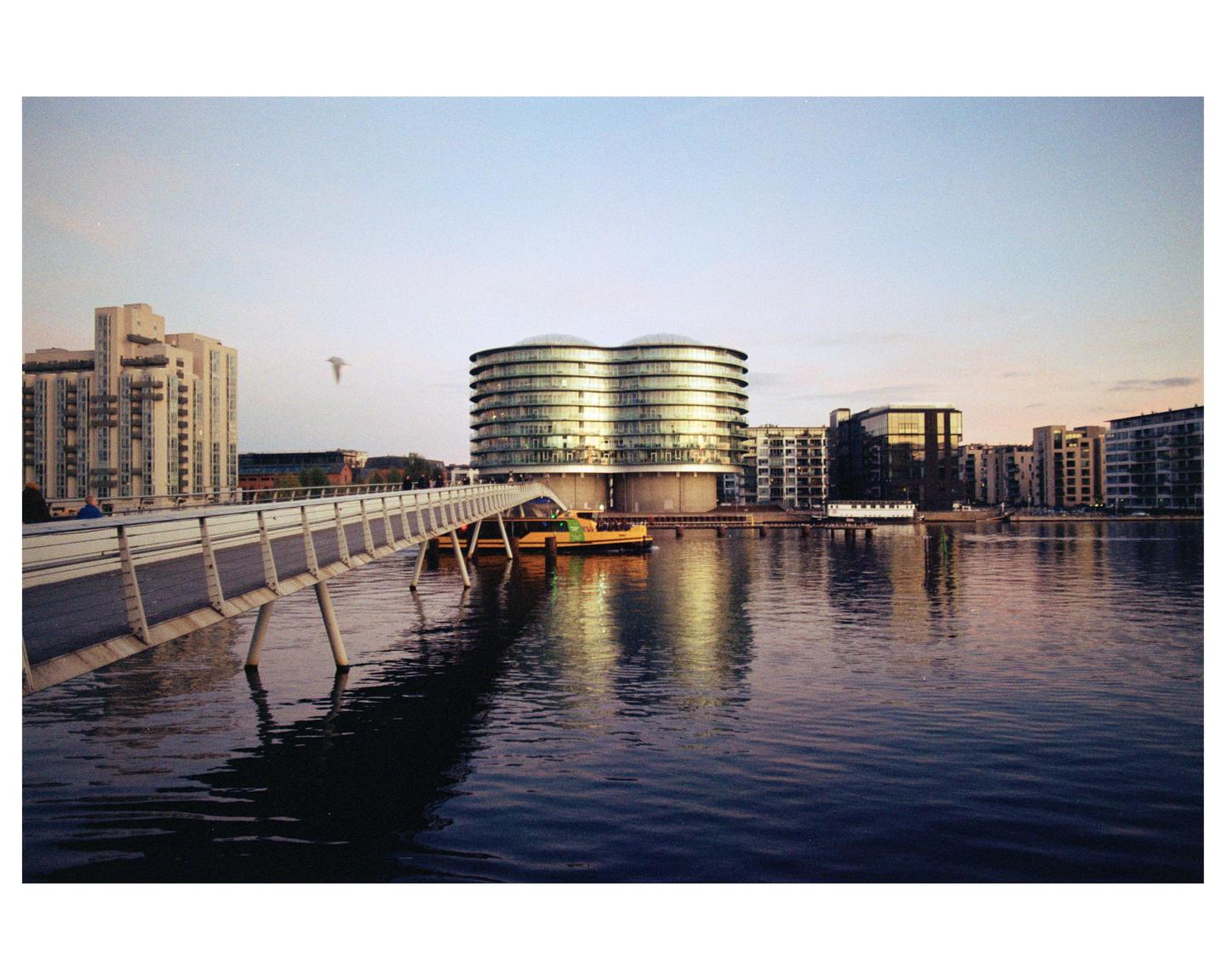 A pedestrian bridge extends over water towards a city skyline with a prominent modern building reflecting the golden hour light.