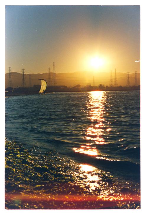 The sun sets over a body of water with a person kitesurfing in the distance and power lines on the horizon.