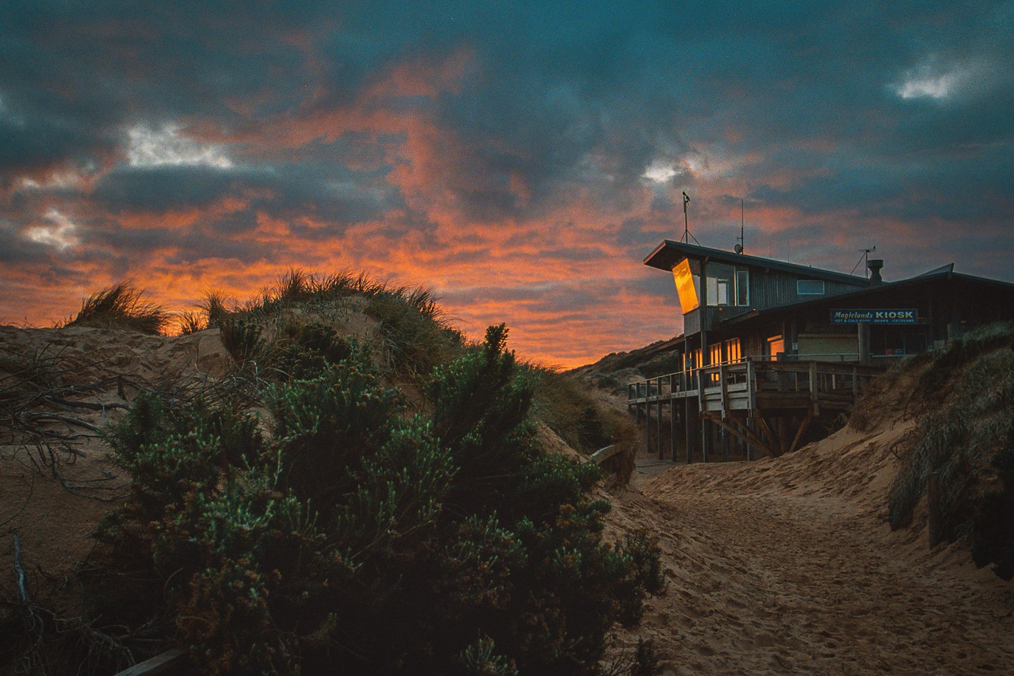 A building with a sign for "Magiclands Kiosk" is nestled in sand dunes under a dramatic sunset sky.