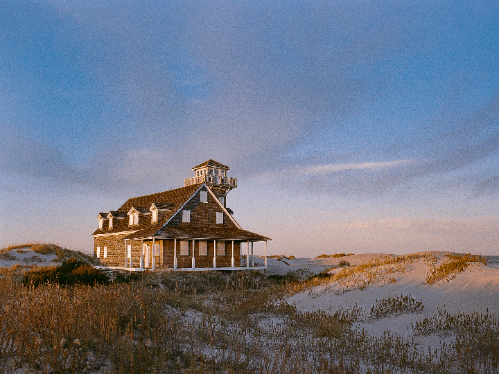 A large house with a tower and porch stands among sand dunes and dry grass under a blue sky.