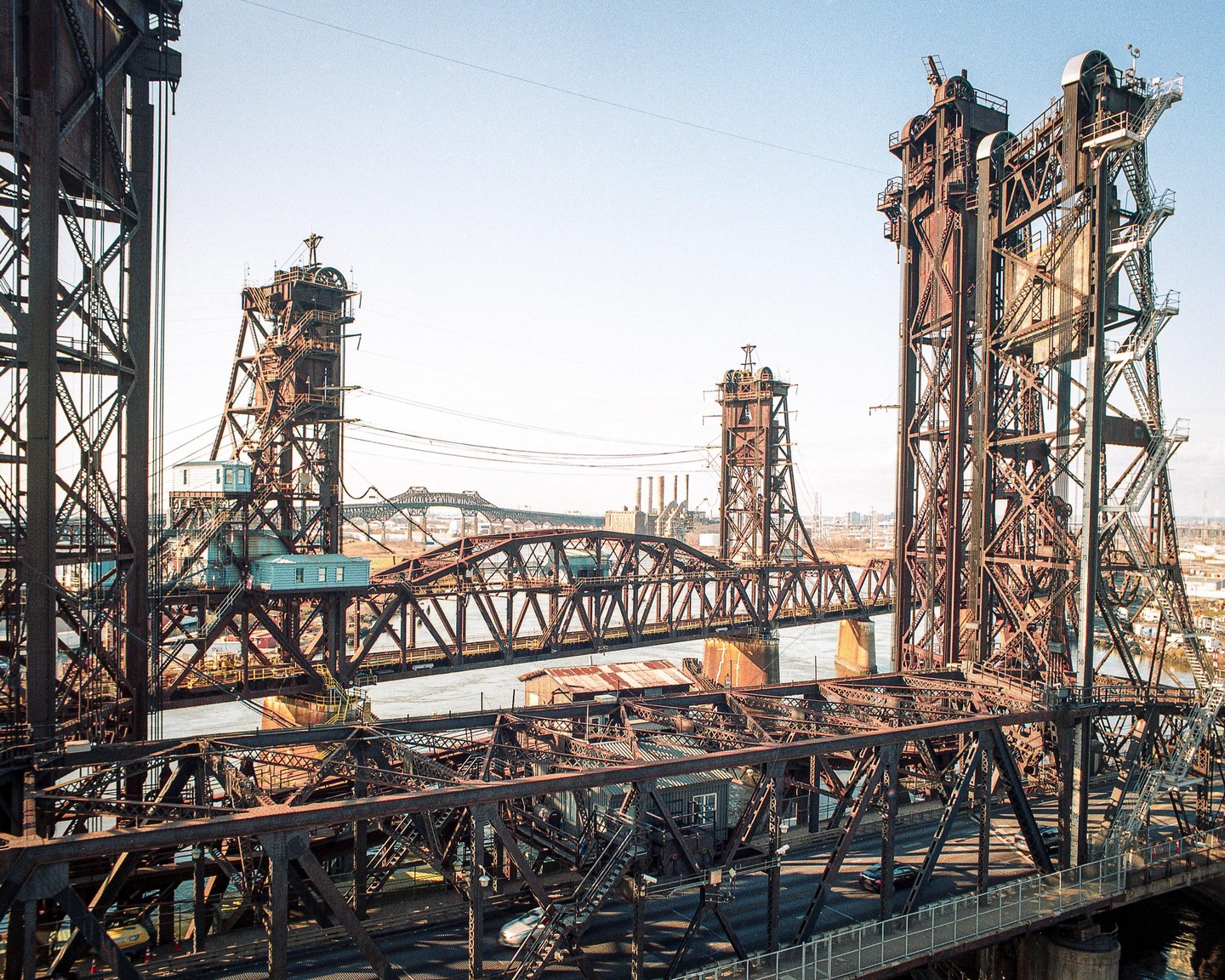 A large metal bridge with multiple spans crosses a body of water, with a city visible in the distance.