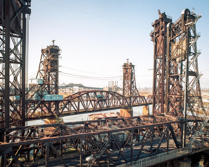 A large metal bridge with multiple spans crosses a body of water, with a city visible in the distance.