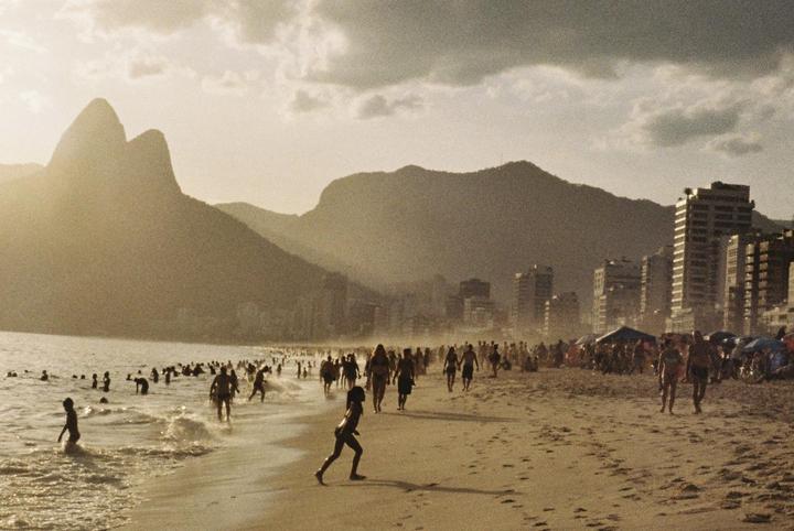 A crowded beach with mountains and buildings in the background is illuminated by the setting sun.