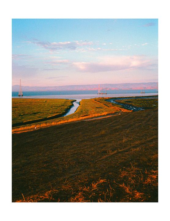 A body of water with a bridge and power lines in the distance is surrounded by fields and a small stream under a colorful sky.