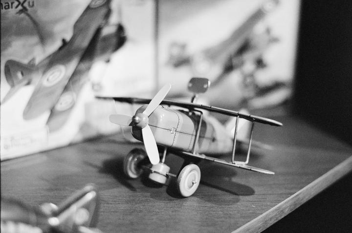 A toy biplane with a propeller and wheels sits on a wooden surface, with other toy planes blurred in the background.
