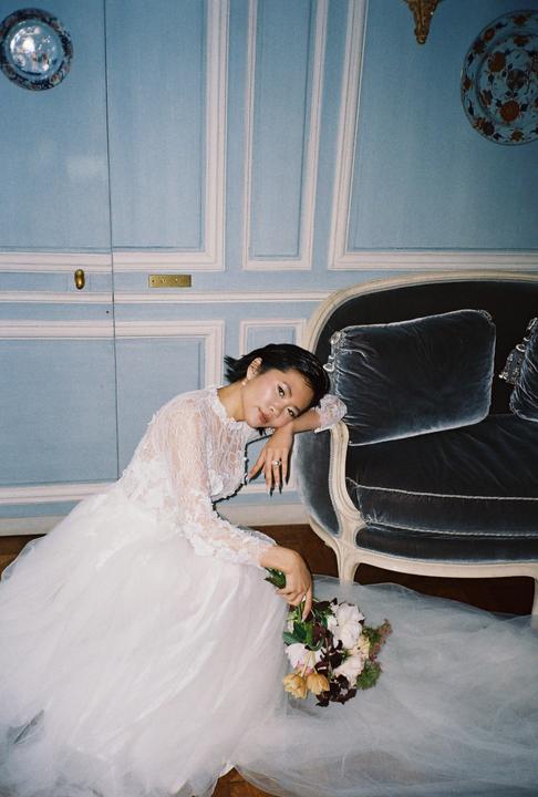 A woman in a white wedding dress is sitting on the floor next to a blue sofa, holding a bouquet of flowers.