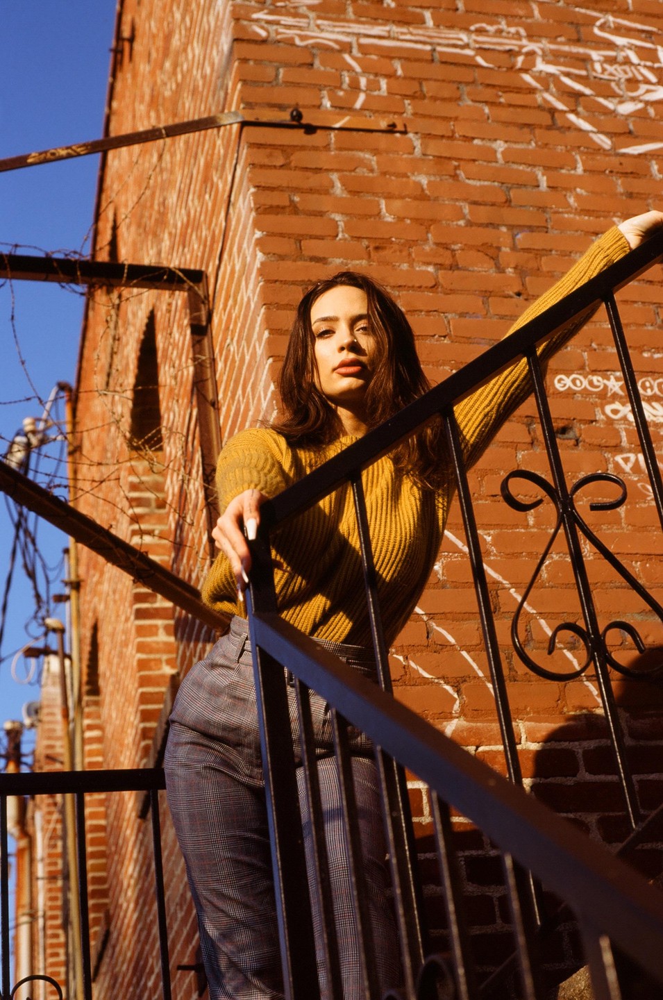 A woman stands on a metal staircase next to a brick building, looking directly at the viewer.