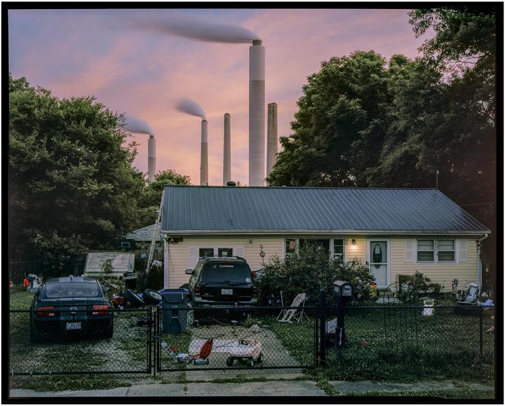A small house with two cars in the driveway is visible behind a fence, with large industrial smokestacks emitting smoke in the background.