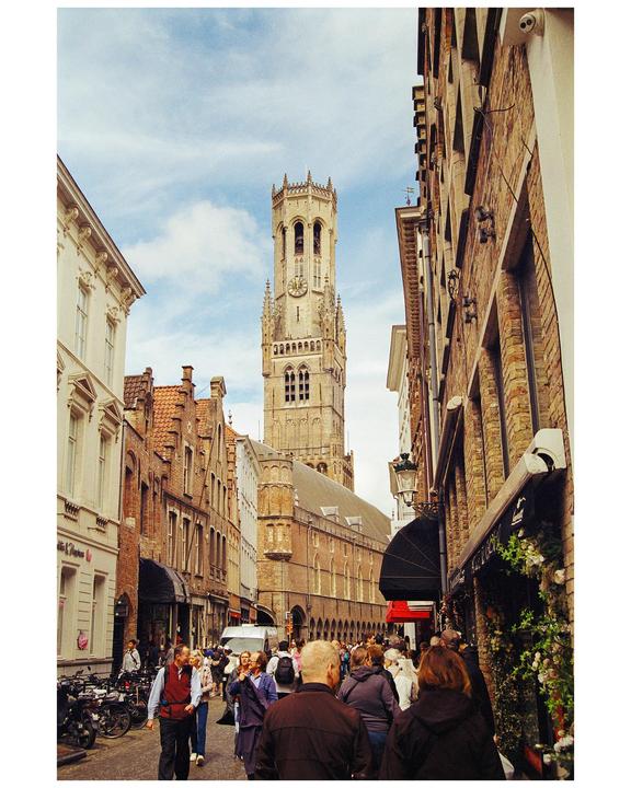 A tall, ornate clock tower rises above a bustling street lined with historic buildings and many people.