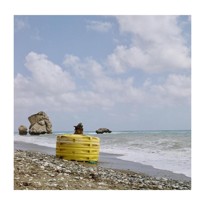 A yellow buoy sits on a rocky beach with the ocean and large rock formations in the background under a cloudy sky.