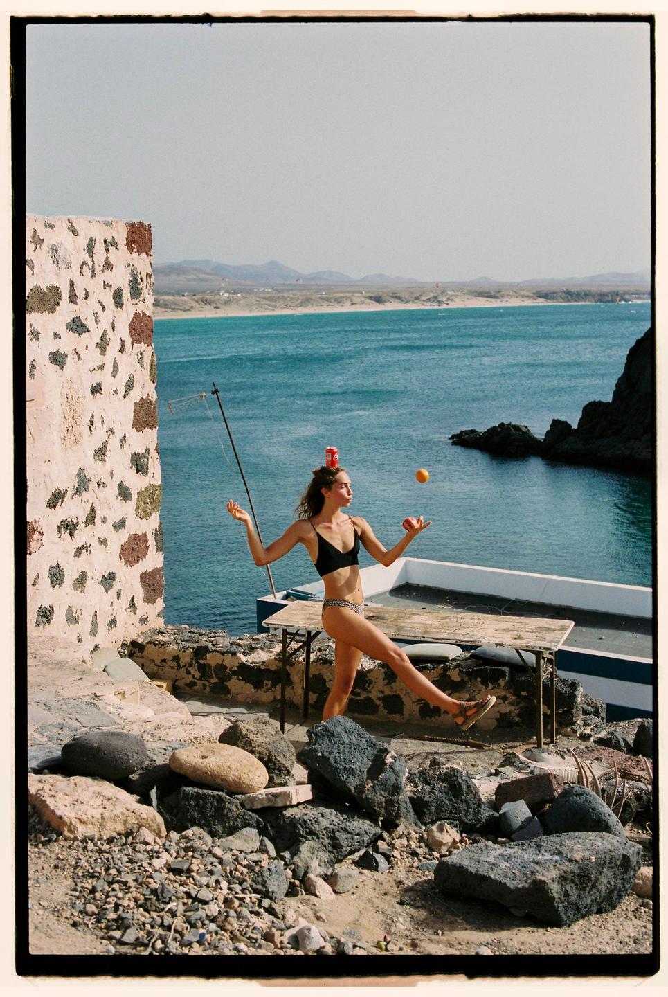 A woman in a bikini balances fruit and a soda can on her head and hands by the sea.