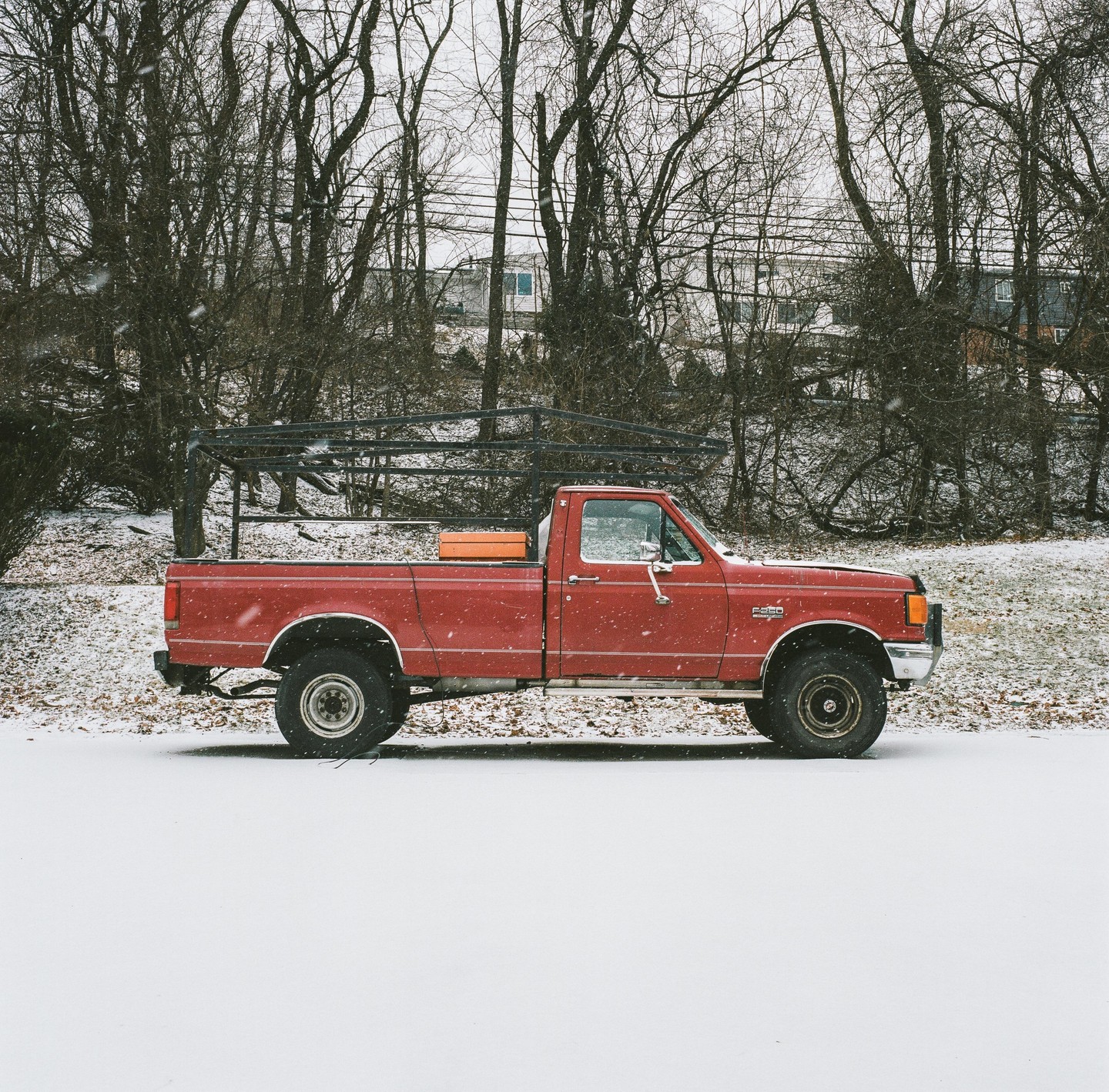 A red pickup truck is parked in a snowy field with bare trees in the background.
