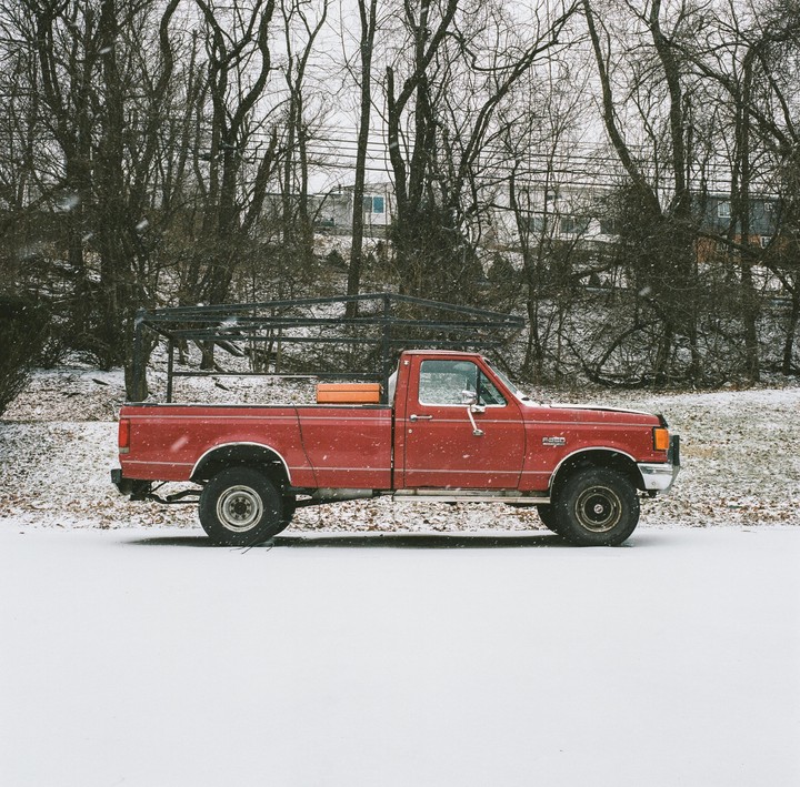 A red pickup truck is parked in a snowy field with bare trees in the background.