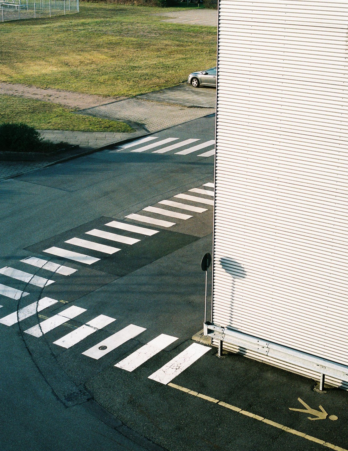 An overhead view shows a crosswalk intersecting with another crosswalk next to a corrugated metal building and a grassy area with a car.