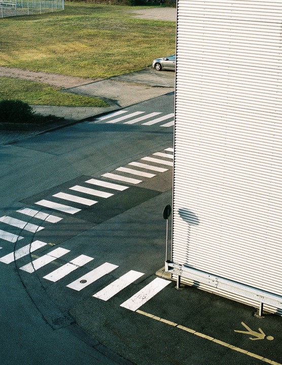 An overhead view shows a crosswalk intersecting with another crosswalk next to a corrugated metal building and a grassy area with a car.