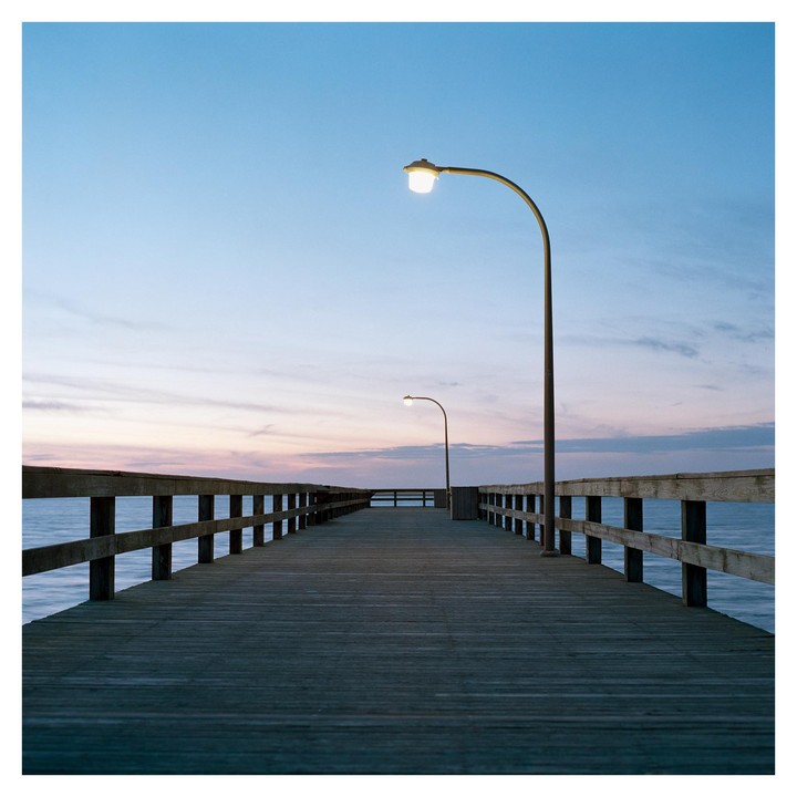 A wooden pier with two streetlights extends into the ocean under a colorful sunset sky.