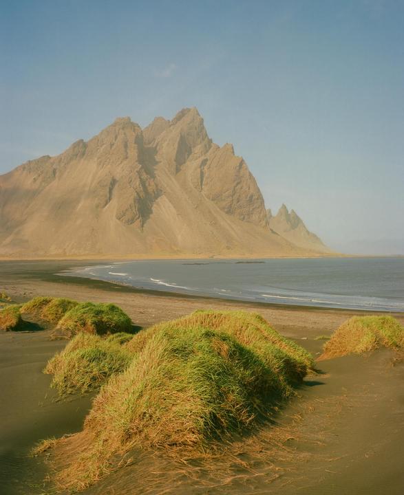 A large mountain range rises behind a black sand beach with green grass tufts in the foreground.