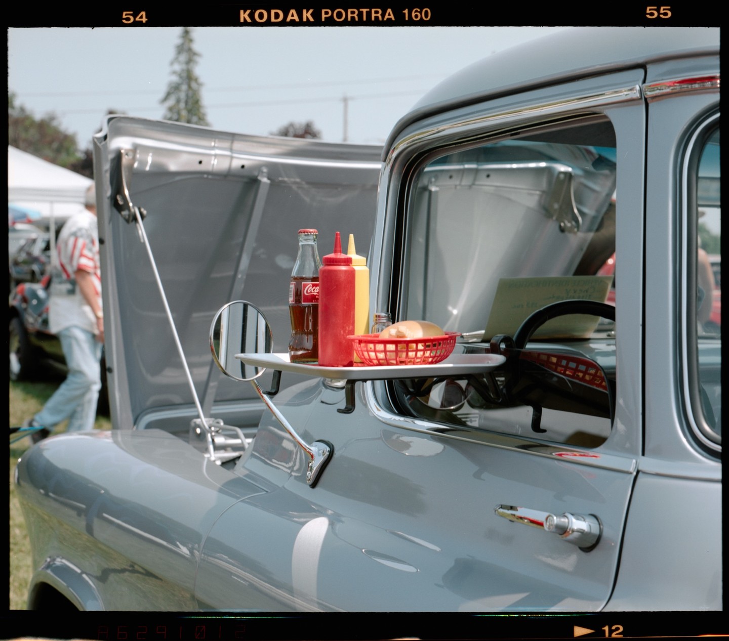 A drive-in tray with a soda, condiments, and a hot dog bun hangs from the window of a vintage truck.