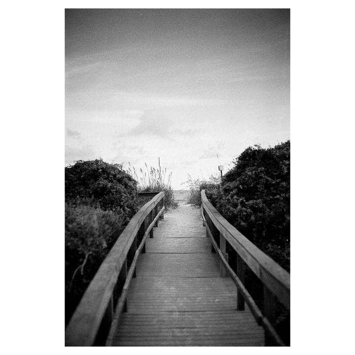 A wooden boardwalk leads to a sandy beach surrounded by bushes and tall grass under a cloudy sky.