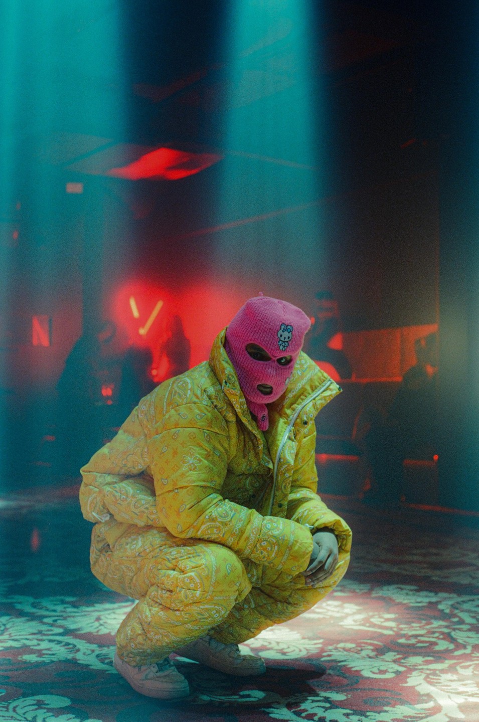 A person in a yellow padded suit and a pink balaclava with a Hello Kitty design crouches on a patterned carpet under blue and red lighting.