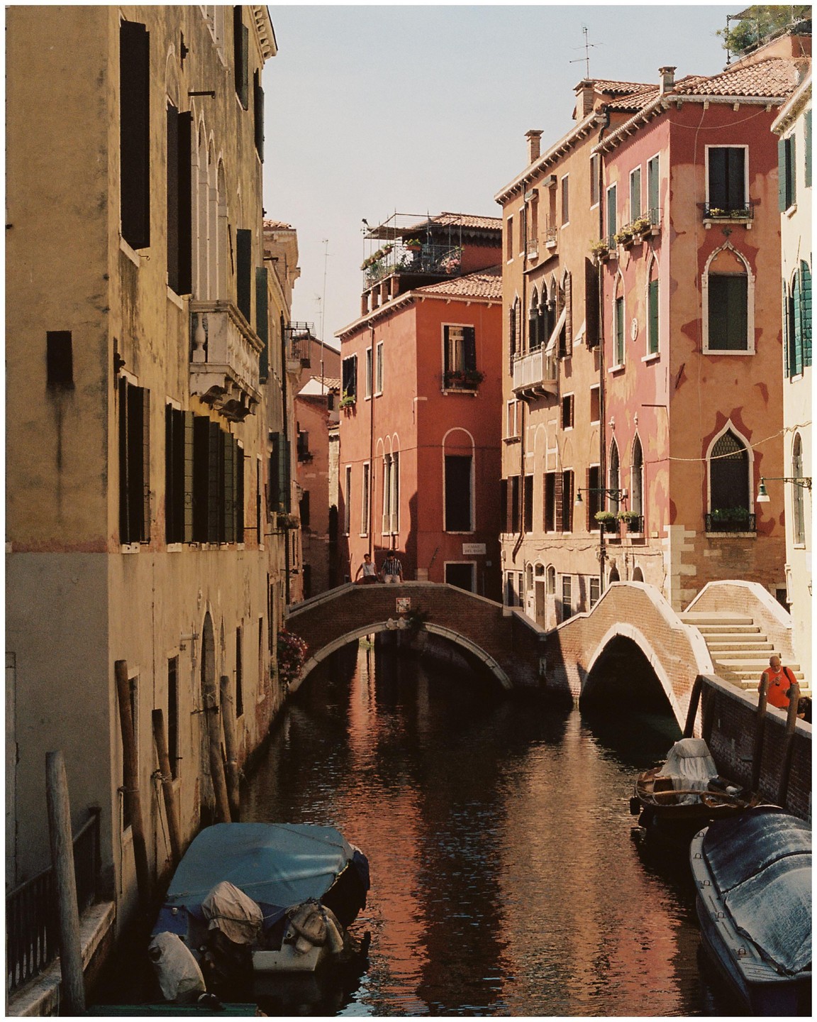 A narrow canal with boats is surrounded by tall buildings and crossed by two arched bridges.