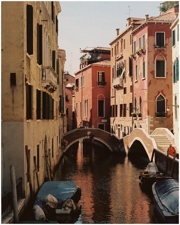 A narrow canal with boats is surrounded by tall buildings and crossed by two arched bridges.