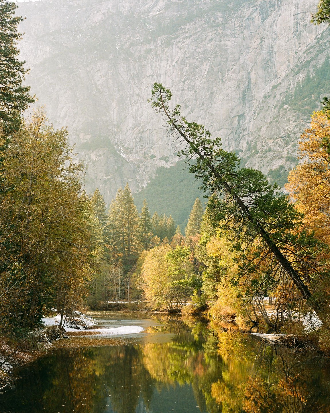 A river reflects autumn trees and a snow-covered bank with a large mountain in the background.