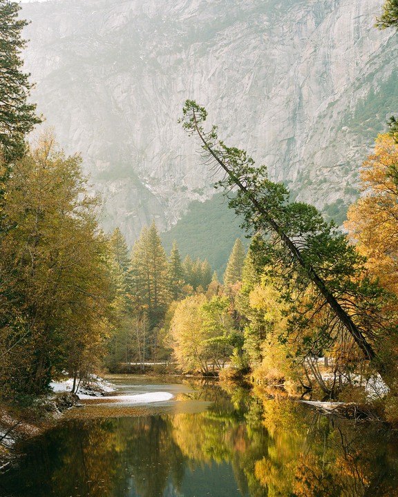A river reflects autumn trees and a snow-covered bank with a large mountain in the background.