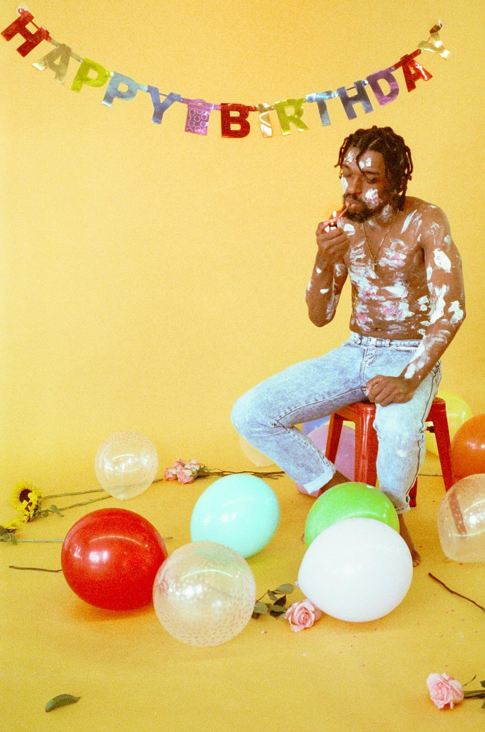 A man covered in cake frosting sits on a red stool, lighting a cigarette under a "Happy Birthday" banner, surrounded by balloons and flowers on a yellow background.