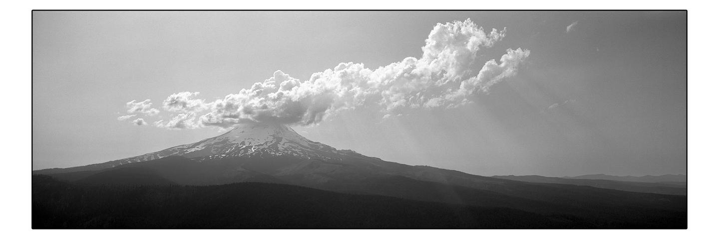 A large cloud formation sits atop a snow-capped mountain with forested hills in the foreground.