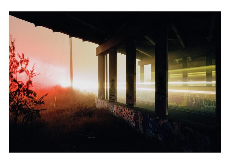 Light trails from vehicles pass under a concrete overpass covered in graffiti during a hazy sunset.