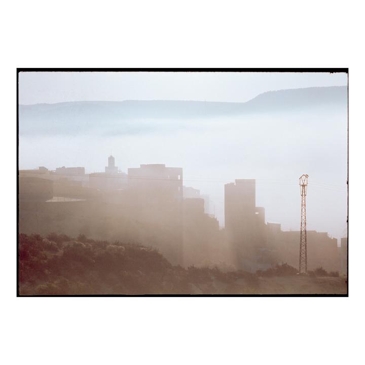 A town with a tall tower is shrouded in fog with mountains in the background.